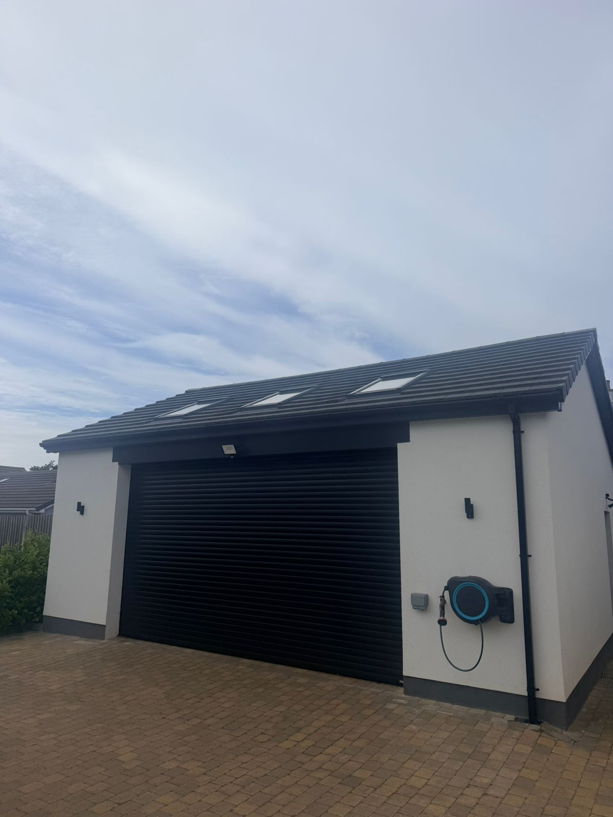 The completed detached triple garage built from the ground up on the Isle of Man by JDM Building Services — white rendered walls, pitched tiled roof with roof windows and a black roller shutter door
