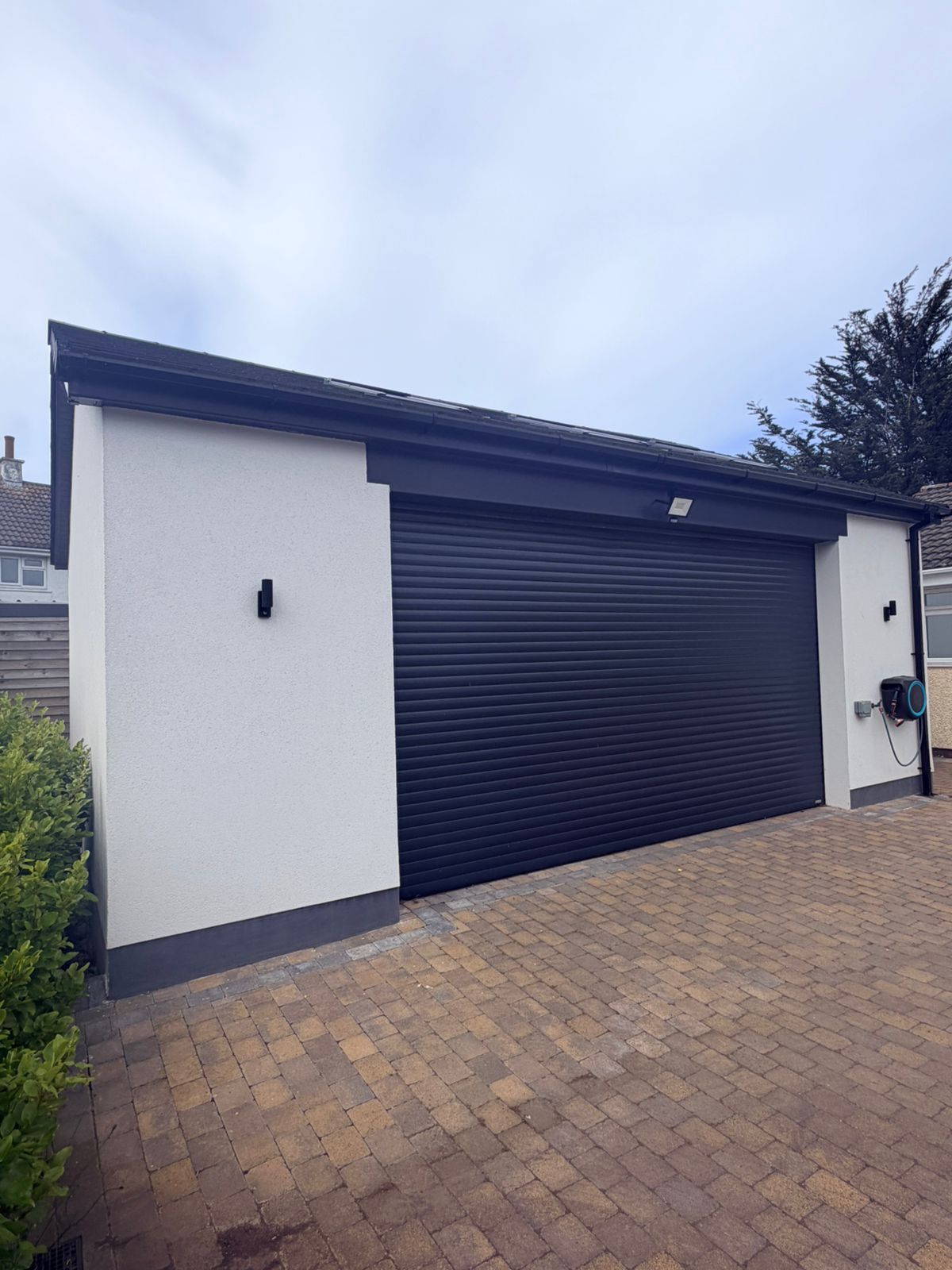 The completed detached triple garage with white rendered walls, a pitched tiled roof and a wide black roller shutter door over a block-paved drive, Isle of Man
