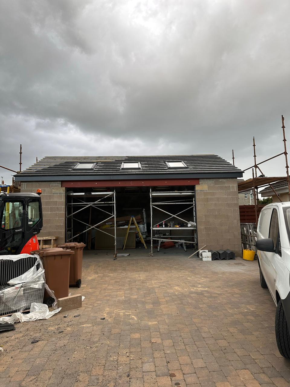 The detached triple garage with its pitched tiled roof and roof windows complete over the blockwork shell, scaffolding still in place inside the openings before render and the roller door, Isle of Man